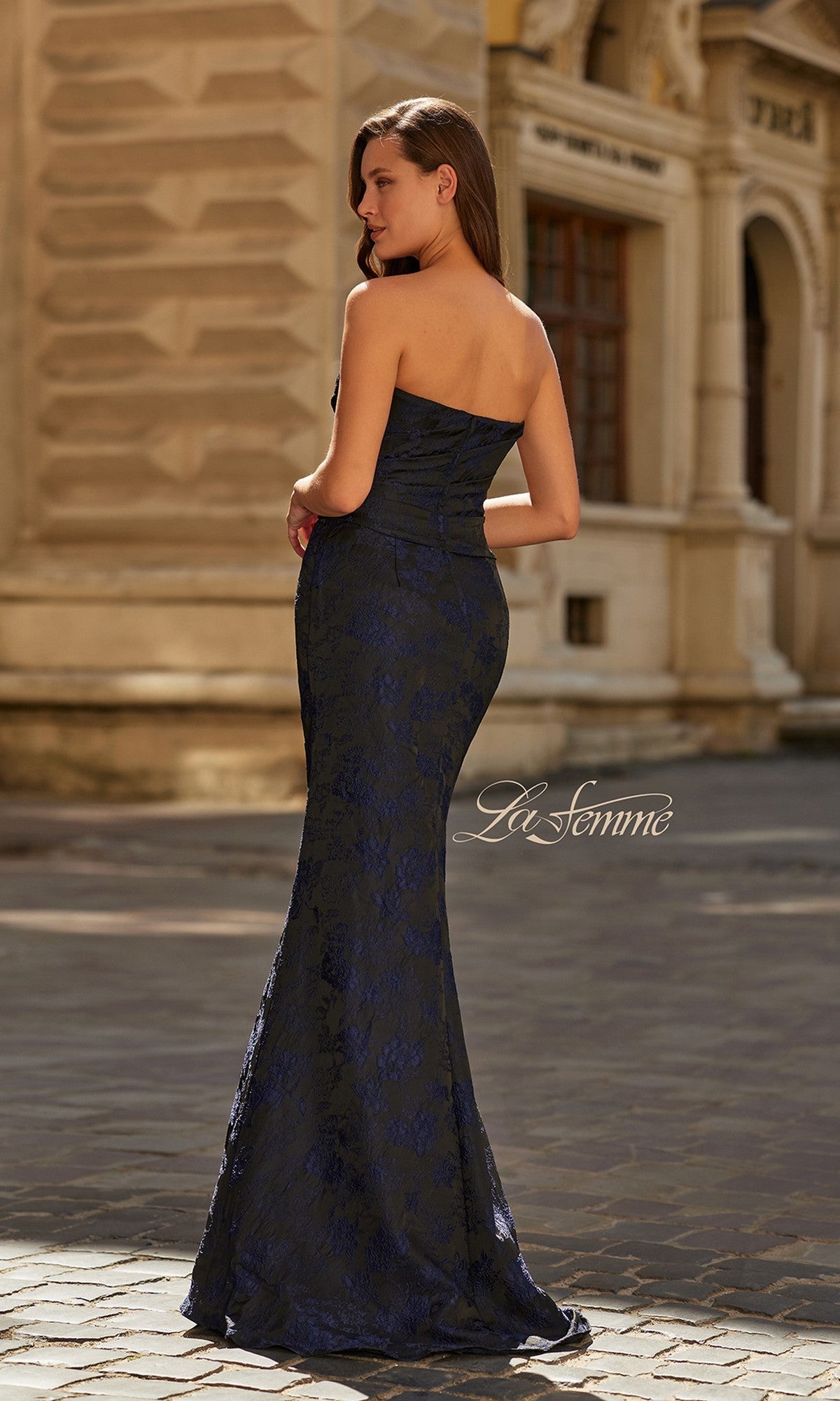A woman wearing the Long Formal Dress: La Femme 33539 stands on a cobblestone street beside an ornate building, facing away from the camera.